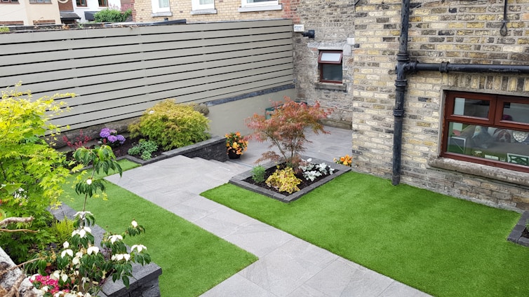 A small, neatly landscaped backyard features green artificial grass and several flower beds with various plants and colorful flowers. The area is enclosed by a brick wall and a wooden fence, providing privacy. A gray stone pathway divides the garden, leading to a door and windows on a brick house facade.