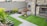 A small, neatly landscaped backyard features green artificial grass and several flower beds with various plants and colorful flowers. The area is enclosed by a brick wall and a wooden fence, providing privacy. A gray stone pathway divides the garden, leading to a door and windows on a brick house facade.
