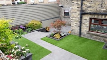 A small, neatly landscaped backyard features green artificial grass and several flower beds with various plants and colorful flowers. The area is enclosed by a brick wall and a wooden fence, providing privacy. A gray stone pathway divides the garden, leading to a door and windows on a brick house facade.