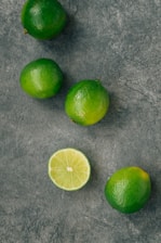 Several whole limes are scattered on a textured gray surface, with one lime cut in half displaying its juicy interior.