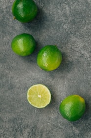 Several whole limes are scattered on a textured gray surface, with one lime cut in half displaying its juicy interior.