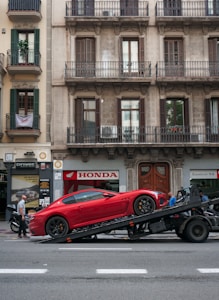 A red sports car is loaded onto a flatbed tow truck parked on a city street. The backdrop features a classic building with ornate balconies and various storefronts including a Honda dealership. Several people are seen around, one walking and another seemingly overseeing the car loading process.