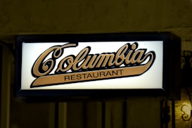An illuminated sign reads 'Columbia Restaurant' in stylized script. The sign has a light background with dark edging, creating a contrast that enhances the visibility of the text.