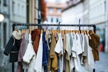 A clothing rack filled with various apparel on wooden hangers is positioned in an outdoor setting. The garments include coats, shirts, sweaters, and cardigans in a range of colors such as black, white, brown, beige, and denim blue. The background features blurred buildings and a street, suggesting a marketplace or an outdoor shopping area.