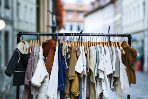 A clothing rack filled with various apparel on wooden hangers is positioned in an outdoor setting. The garments include coats, shirts, sweaters, and cardigans in a range of colors such as black, white, brown, beige, and denim blue. The background features blurred buildings and a street, suggesting a marketplace or an outdoor shopping area.