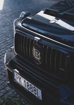 A close-up of the front end of a black luxury car with a prominent 'B' logo on the grille and a license plate reading 'PK 1888L'. The car is parked on a cobblestone surface, with reflections visible on the vehicle's sleek, polished surface.