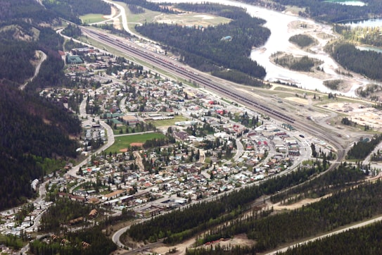 An aerial view of a small town surrounded by a dense forest. Roads and residential buildings, along with an expanse of railway tracks, are clearly visible. The town is bordered by a winding river on one side, and verdant forested hills on the other. The landscape features a mixture of urban and natural elements.