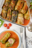 A dish of stuffed cabbage rolls covered in tomato sauce is displayed in a glass baking dish, alongside a plated portion showing two rolls with one cut open, revealing a meat filling. Fresh cherry tomatoes and silverware are arranged neatly nearby.