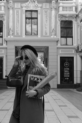 A stylish person in a long coat and beret holds a magazine and a baguette while adjusting sunglasses. The background features an ornate building facade with intricate architectural details, including decorated columns and arched windows.