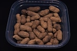 A clear plastic container holds several unshelled peanuts against a dark background. The peanuts are arranged in a random pile, showcasing their textured, rough shells with a light brown color.