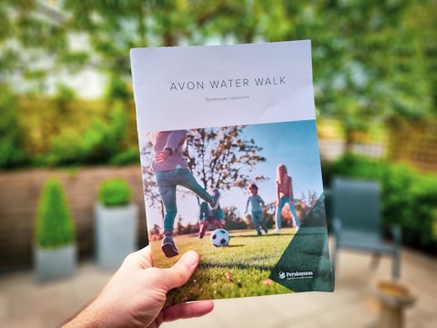 A person is holding a brochure titled 'AVON WATER WALK' with an image of children playing soccer on a field. The background is a blurred patio with greenery and outdoor furniture.