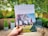 A person is holding a brochure titled 'AVON WATER WALK' with an image of children playing soccer on a field. The background is a blurred patio with greenery and outdoor furniture.