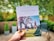 A person is holding a brochure titled 'AVON WATER WALK' with an image of children playing soccer on a field. The background is a blurred patio with greenery and outdoor furniture.