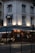 A corner café with a classic European architectural style. The building is white with dark blue awnings, typical of many quaint cafés in urban settings. The warm interior lighting contrasts with the dim outdoor light, suggesting early evening. Chairs and tables are set outside, with a couple of patrons possibly enjoying a meal or drink.