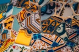 A close-up of intricately patterned mosaic art made of various colorful ceramic or tile pieces. The fragments depict a mix of geometric and floral designs, featuring an array of shapes and patterns. The tiles are mostly seamless with complementary and contrasting colors that create a visually stimulating composition.