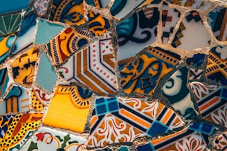 A close-up of intricately patterned mosaic art made of various colorful ceramic or tile pieces. The fragments depict a mix of geometric and floral designs, featuring an array of shapes and patterns. The tiles are mostly seamless with complementary and contrasting colors that create a visually stimulating composition.