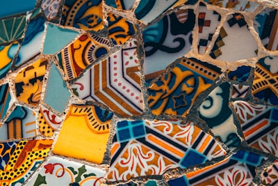 A close-up of intricately patterned mosaic art made of various colorful ceramic or tile pieces. The fragments depict a mix of geometric and floral designs, featuring an array of shapes and patterns. The tiles are mostly seamless with complementary and contrasting colors that create a visually stimulating composition.