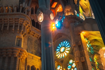 Intricate architectural details of a grand building interior are portrayed, highlighting large stained glass windows that cast colorful light patterns across stone walls and columns. The style features elaborate carvings and geometric shapes, with vibrant blue, yellow, and orange hues illuminated by the sunlight.