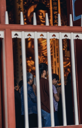 A group of people stands in prayer in front of a large golden Buddha statue inside a temple, viewed through vertical bars. The atmosphere is solemn and respectful, with individuals holding their hands together in reverence.