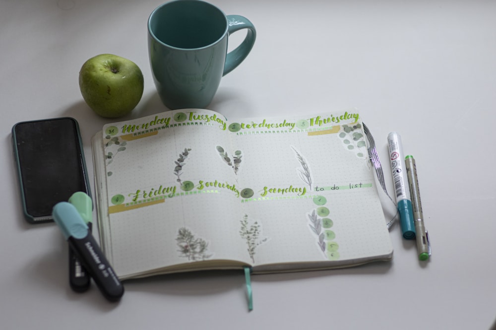 Tasse en céramique verte à côté du stylo à clic noir sur livre blanc et vert