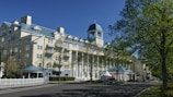 A large, elegant hotel with a light yellow facade and white columns stands prominently against a clear blue sky. The building features multiple windows and a distinctive tower with a clock. The foreground includes a well-maintained road lined with green trees, casting shadows on the pavement.