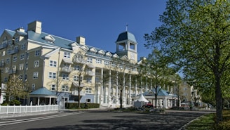 A large, elegant hotel with a light yellow facade and white columns stands prominently against a clear blue sky. The building features multiple windows and a distinctive tower with a clock. The foreground includes a well-maintained road lined with green trees, casting shadows on the pavement.