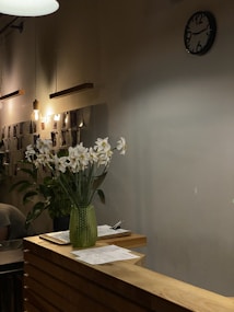 A cozy interior with warm lighting, featuring a wooden counter adorned with white flowers in a green glass vase. On the counter, there are papers and a pen. The background includes a wall-mounted clock and hanging light bulbs, with photos or documents displayed on the wall.
