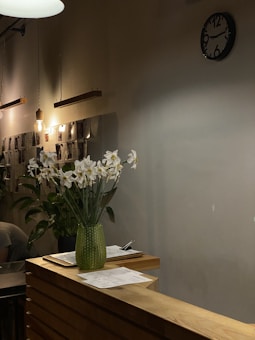 A cozy interior with warm lighting, featuring a wooden counter adorned with white flowers in a green glass vase. On the counter, there are papers and a pen. The background includes a wall-mounted clock and hanging light bulbs, with photos or documents displayed on the wall.