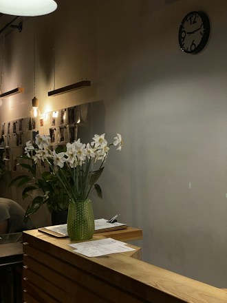 A cozy interior with warm lighting, featuring a wooden counter adorned with white flowers in a green glass vase. On the counter, there are papers and a pen. The background includes a wall-mounted clock and hanging light bulbs, with photos or documents displayed on the wall.