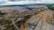 An expansive open-pit mine stretches across a wide landscape with large trenches and piles of earth. Sparse vegetation can be seen at the edges, and heavy machinery is visible within the excavation area. The horizon is lined with hills and patches of green, indicating surrounding forest or fields.