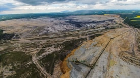 An expansive open-pit mine stretches across a wide landscape with large trenches and piles of earth. Sparse vegetation can be seen at the edges, and heavy machinery is visible within the excavation area. The horizon is lined with hills and patches of green, indicating surrounding forest or fields.