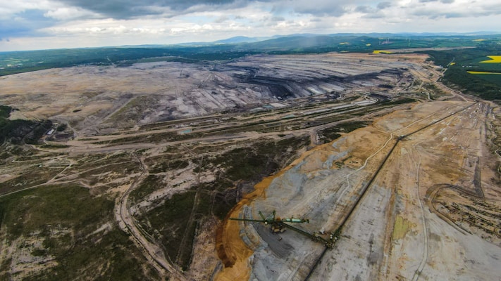 An expansive open-pit mine stretches across a wide landscape with large trenches and piles of earth. Sparse vegetation can be seen at the edges, and heavy machinery is visible within the excavation area. The horizon is lined with hills and patches of green, indicating surrounding forest or fields.