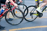 Two cyclists riding closely together on a street, one wearing a yellow and blue outfit with white shoes, and the other in a black and red outfit with blue and black shoes. Both are on road bicycles with visible brand names. There is a water bottle attached to one of the bicycles.