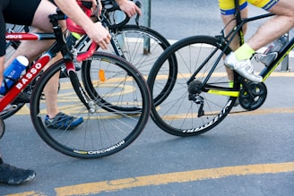 Two cyclists riding closely together on a street, one wearing a yellow and blue outfit with white shoes, and the other in a black and red outfit with blue and black shoes. Both are on road bicycles with visible brand names. There is a water bottle attached to one of the bicycles.