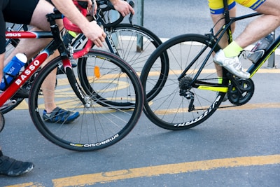 Two cyclists riding closely together on a street, one wearing a yellow and blue outfit with white shoes, and the other in a black and red outfit with blue and black shoes. Both are on road bicycles with visible brand names. There is a water bottle attached to one of the bicycles.