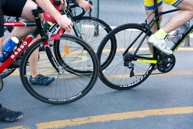 Two cyclists riding closely together on a street, one wearing a yellow and blue outfit with white shoes, and the other in a black and red outfit with blue and black shoes. Both are on road bicycles with visible brand names. There is a water bottle attached to one of the bicycles.