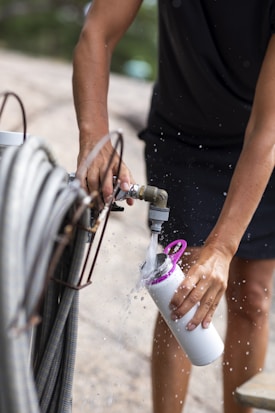 A person fills a white water bottle using an outdoor hose. Water is seen splashing as it flows into the bottle. The person is wearing a black shirt and their hands are visible in the process.