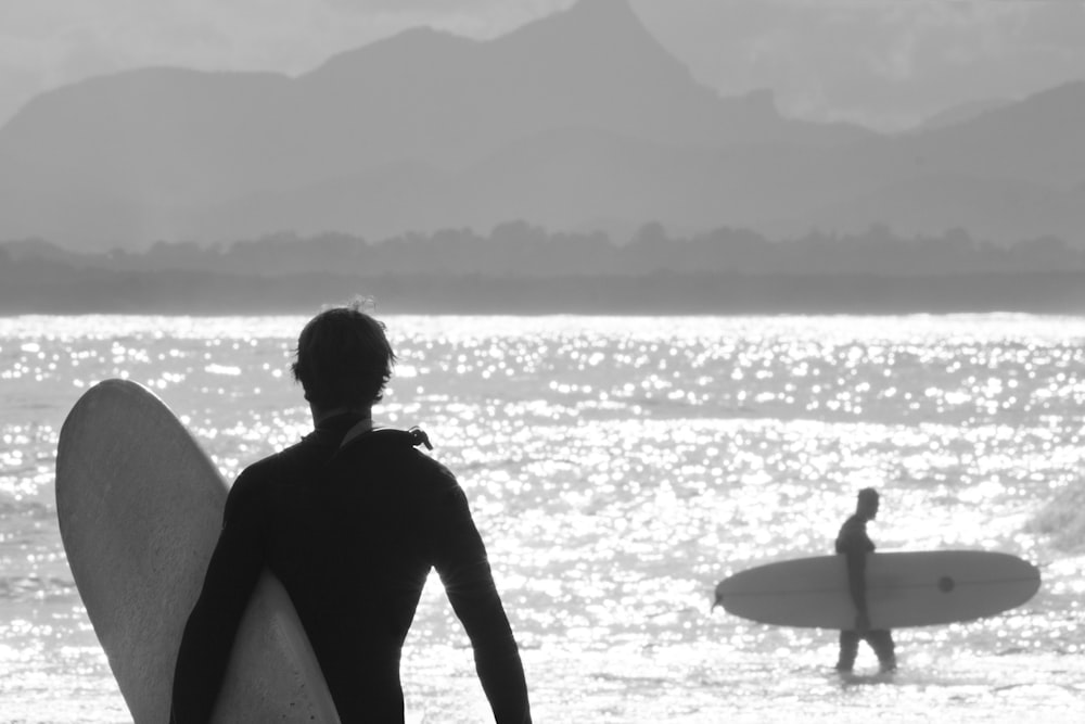 Photo en niveaux de gris d’un homme en combinaison noire tenant une planche de surf