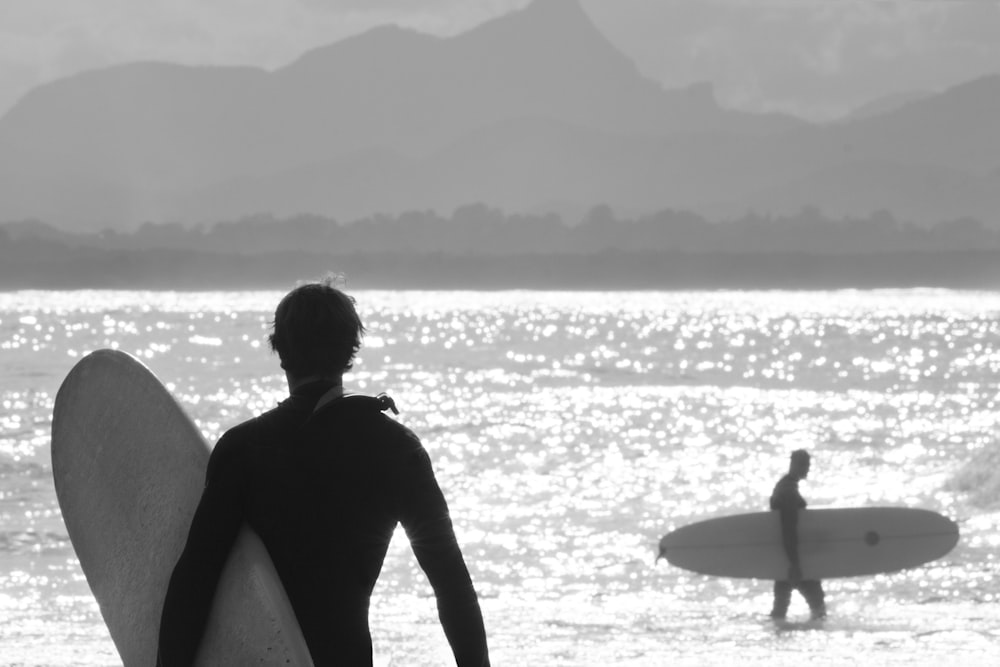 grayscale photo of man in black wet suit holding surfboard