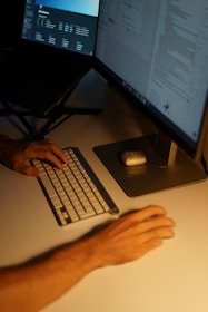 A dimly lit workspace featuring a person typing on a compact keyboard with one hand, while the other hand is placed on the table near a mouse. Two computer monitors display various digital content, creating an atmosphere of focus and concentration.