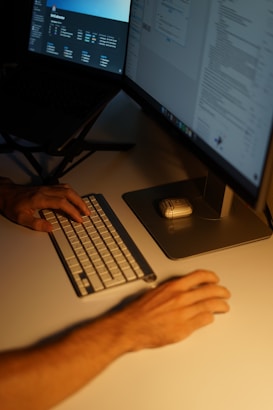 A dimly lit workspace featuring a person typing on a compact keyboard with one hand, while the other hand is placed on the table near a mouse. Two computer monitors display various digital content, creating an atmosphere of focus and concentration.