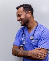 A person with short dark hair is smiling and wearing a blue medical scrub with a stethoscope around their neck. They have tattoos on their forearms and are standing against a plain background with arms crossed.