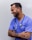 A person with short dark hair is smiling and wearing a blue medical scrub with a stethoscope around their neck. They have tattoos on their forearms and are standing against a plain background with arms crossed.