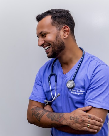 A person with short dark hair is smiling and wearing a blue medical scrub with a stethoscope around their neck. They have tattoos on their forearms and are standing against a plain background with arms crossed.