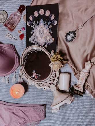 The image contains a variety of objects arranged on a fabric surface, including a decorative black book with moon phases and intricate designs, a vintage-style red perfume bottle, a round mirror with an ornate frame and a small butterfly decoration, a purple bra and panties set, assorted small heart-shaped gemstones, a lit candle, a small statue resembling classical sculpture, and an ornate cameo brooch on a chain.