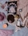 The image contains a variety of objects arranged on a fabric surface, including a decorative black book with moon phases and intricate designs, a vintage-style red perfume bottle, a round mirror with an ornate frame and a small butterfly decoration, a purple bra and panties set, assorted small heart-shaped gemstones, a lit candle, a small statue resembling classical sculpture, and an ornate cameo brooch on a chain.