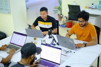 Several people are seated around a table with laptops, engaged in work or study. The environment appears to be a modern office or co-working space, with white tables, chairs, and various office supplies. The individuals are focused on their screens, suggesting a collaborative or tech-related setting.
