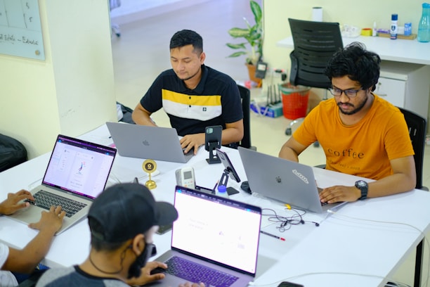 Several people are seated around a table with laptops, engaged in work or study. The environment appears to be a modern office or co-working space, with white tables, chairs, and various office supplies. The individuals are focused on their screens, suggesting a collaborative or tech-related setting.