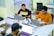 Several people are seated around a table with laptops, engaged in work or study. The environment appears to be a modern office or co-working space, with white tables, chairs, and various office supplies. The individuals are focused on their screens, suggesting a collaborative or tech-related setting.