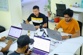 Several people are seated around a table with laptops, engaged in work or study. The environment appears to be a modern office or co-working space, with white tables, chairs, and various office supplies. The individuals are focused on their screens, suggesting a collaborative or tech-related setting.